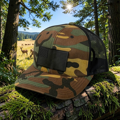 Crafted Merch camouflage trucker hat with a black monochromatic American flag patch, set on a mossy log in a forest.
