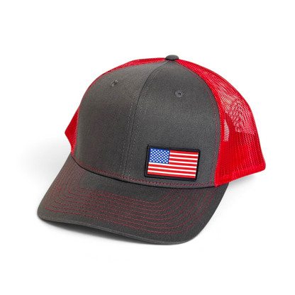 Crafted Merch American flag PVC patch trucker hat in charcoal grey with a vibrant red mesh back and red stitching.